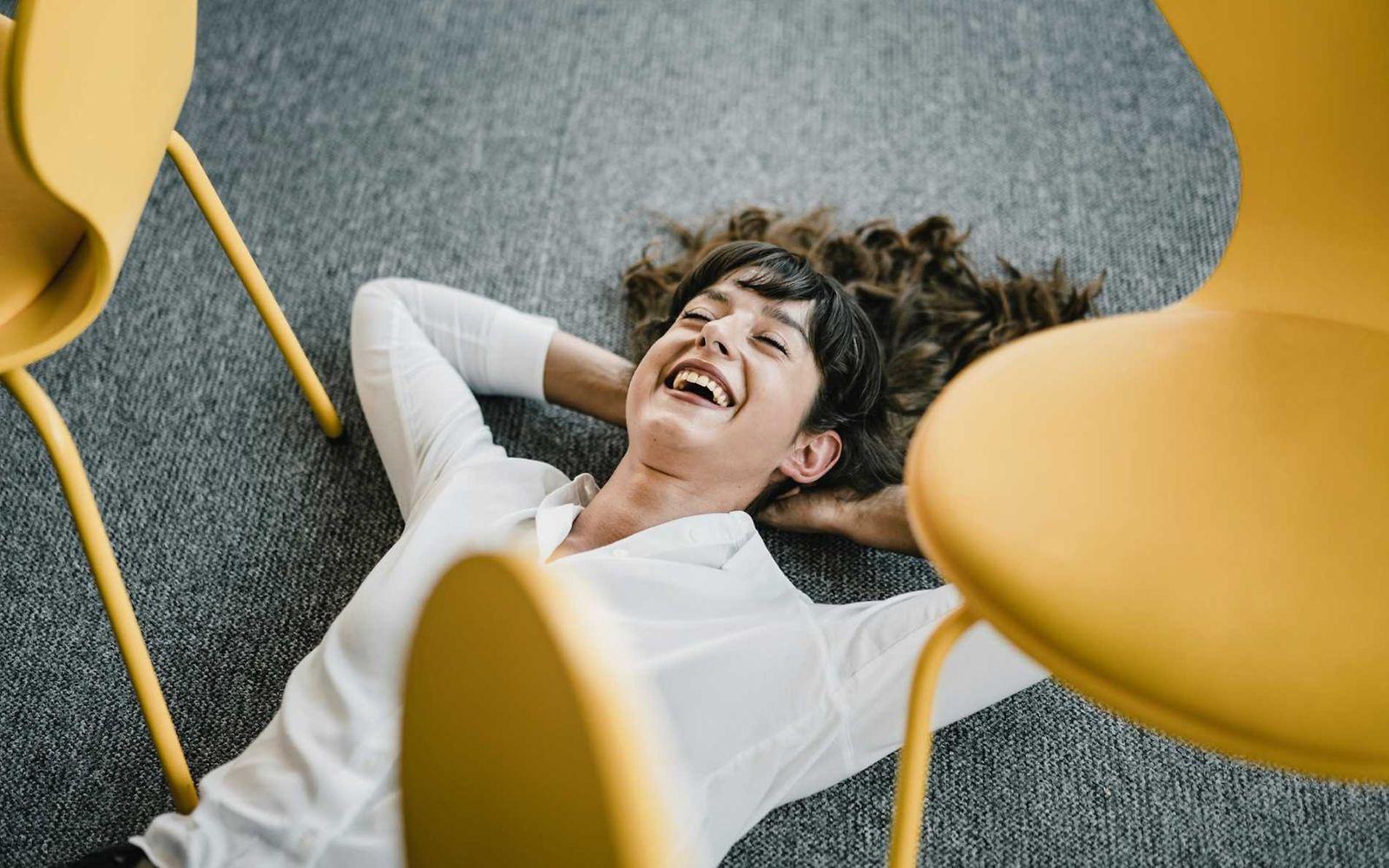 Bild: Frau mit weißer Bluse liegt lächelnd auf dem Fußboden im Büro und wirkt entspannt.