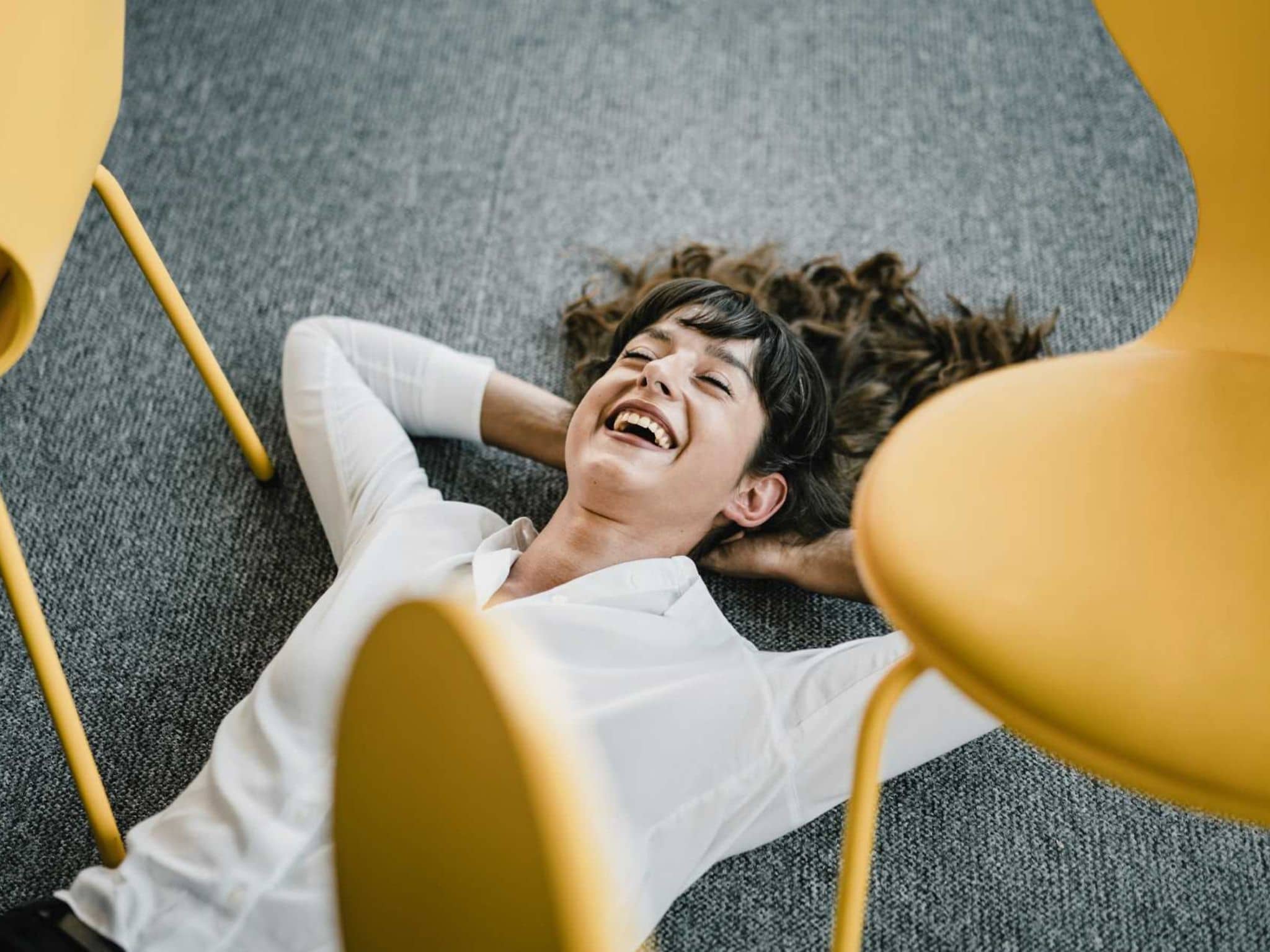 Bild: Frau mit weißer Bluse liegt lächelnd auf dem Fußboden im Büro und wirkt entspannt.