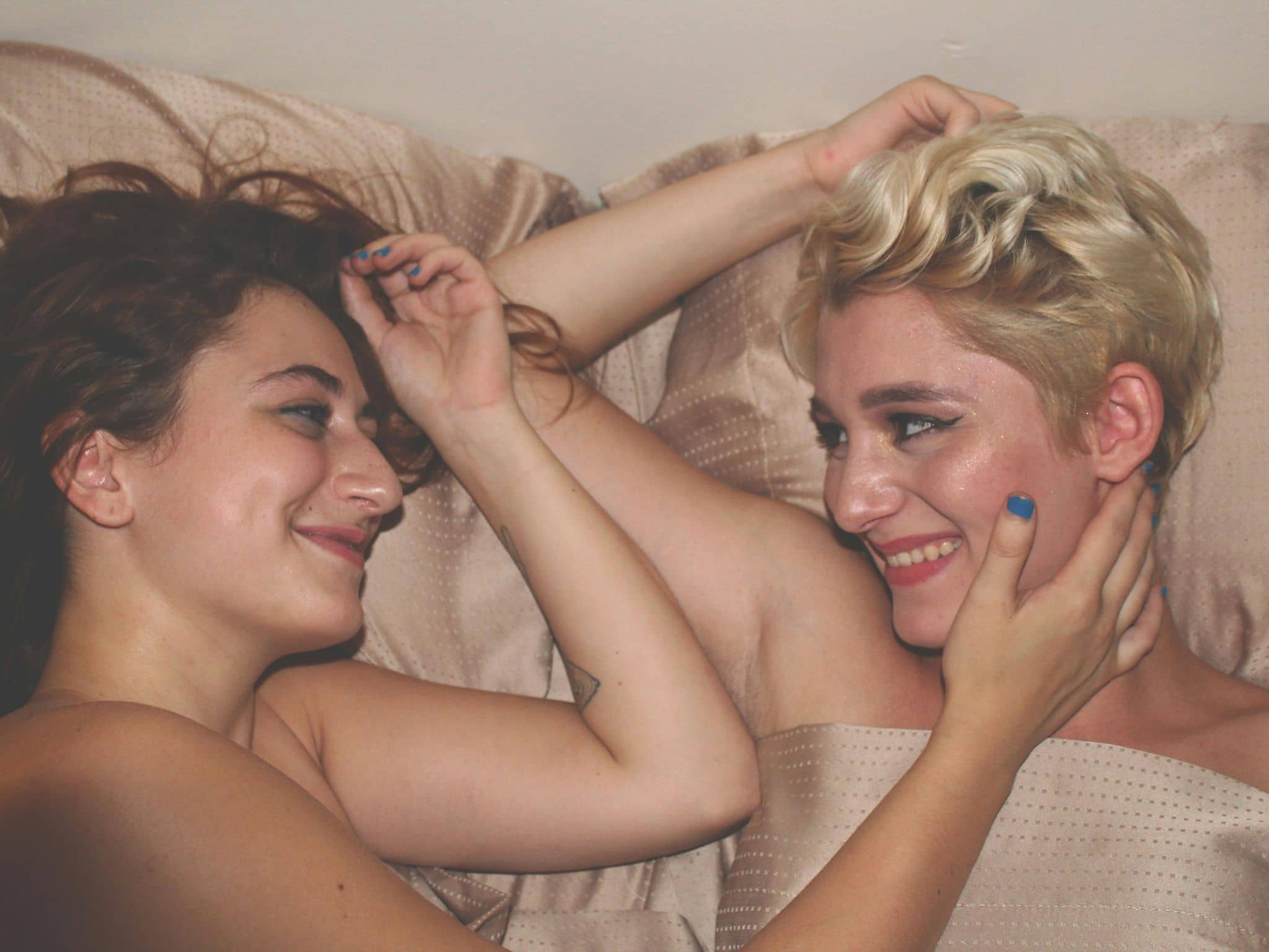 Zwei Frauen liegen nackt im Bett und lächeln sich an.