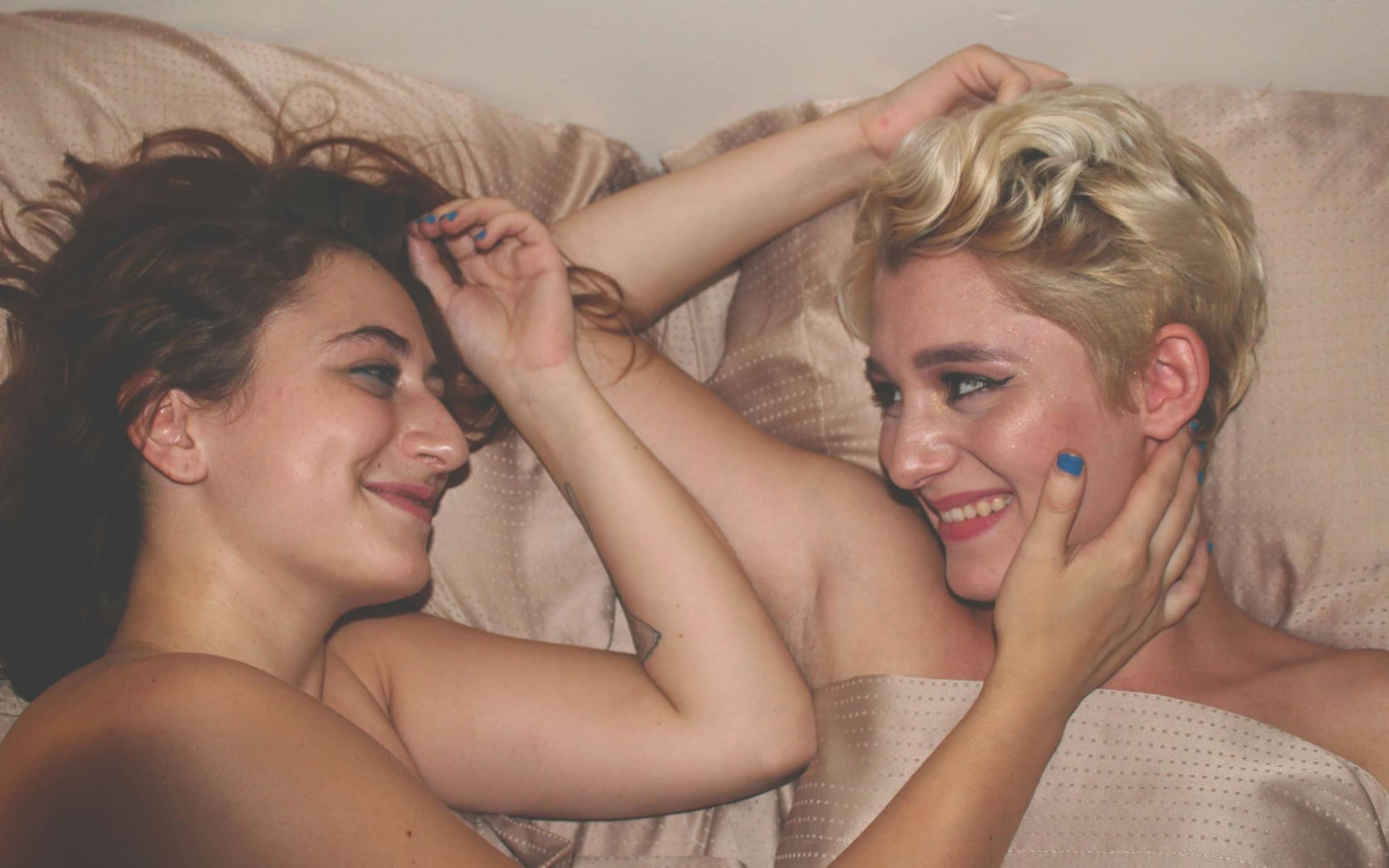 Zwei Frauen liegen nackt im Bett und lächeln sich an.