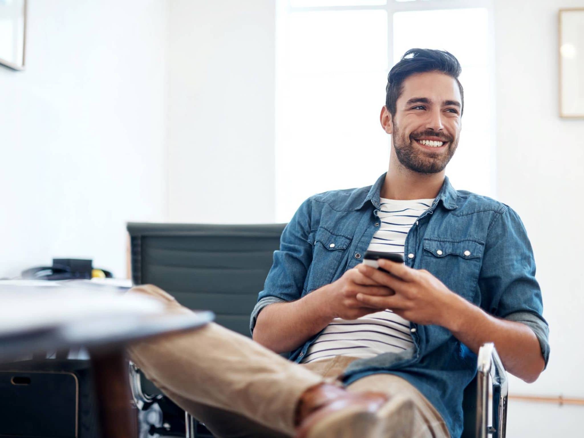 Nebenberuflich selbstständig in der Krankenversicherung: Junger Mann sitzt auf Bürostuhl und hält Handy in der Hand.