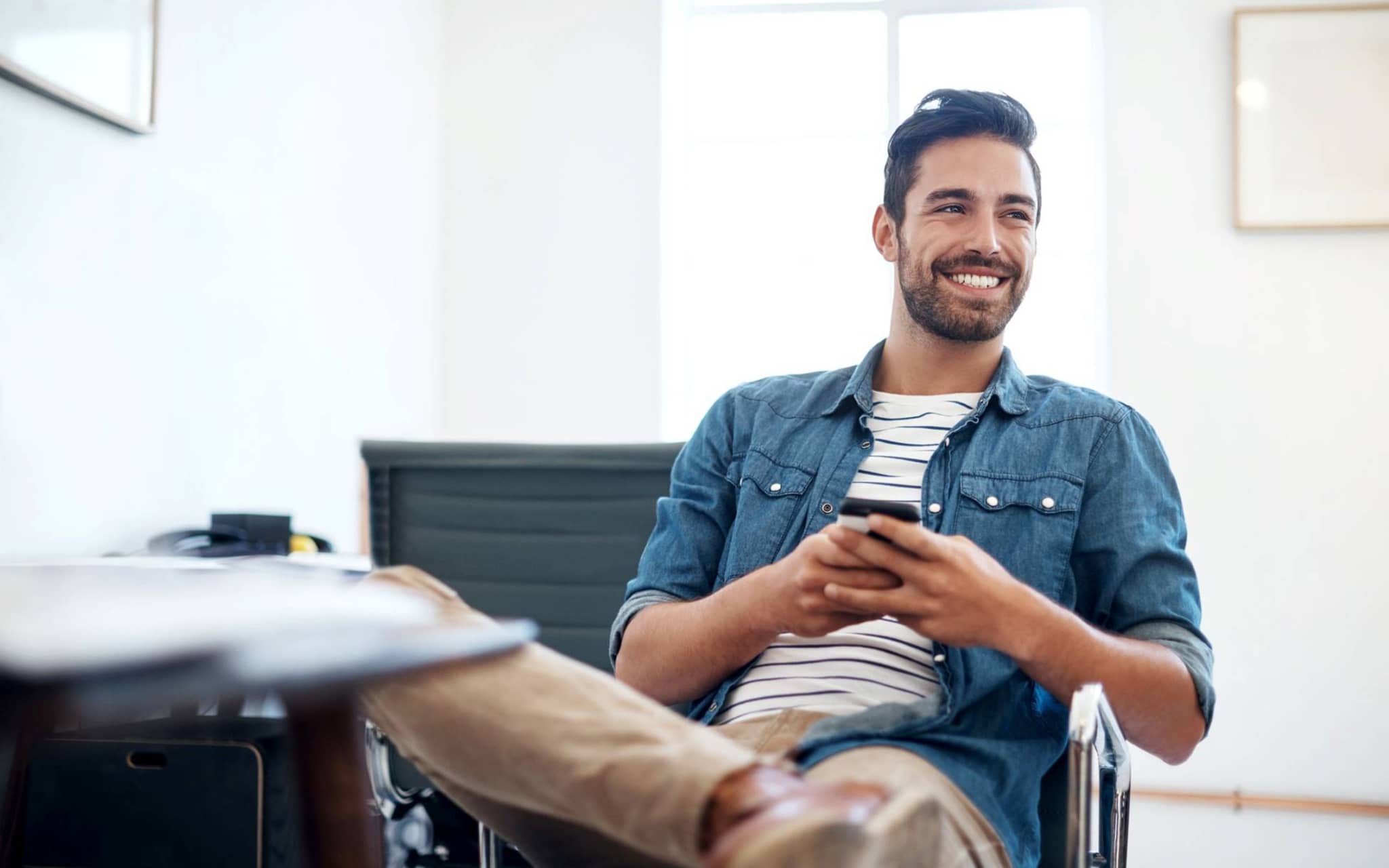 Nebenberuflich selbstständig in der Krankenversicherung: Junger Mann sitzt auf Bürostuhl und hält Handy in der Hand.
