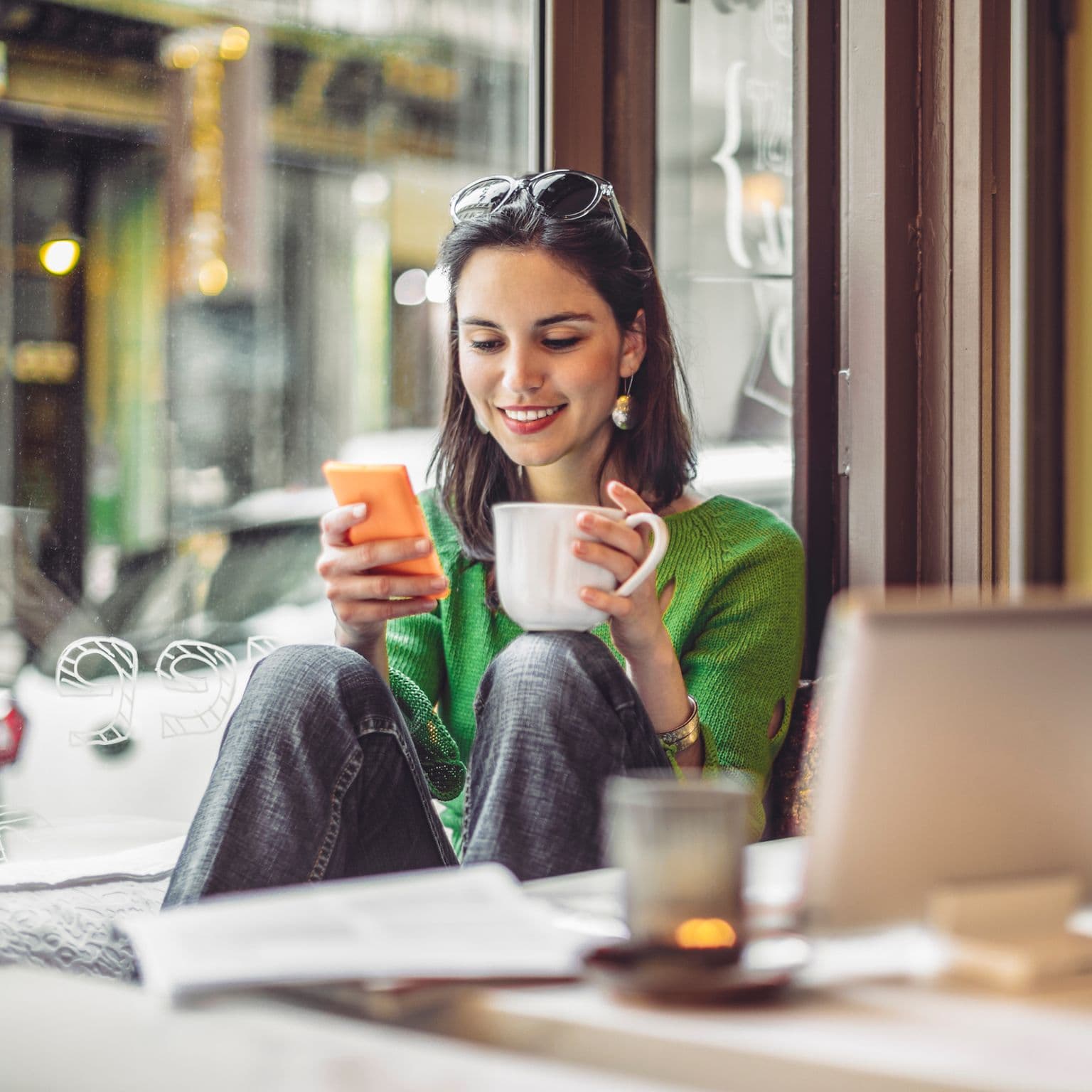 Frau sitzt im Café, guckt lachend in ihr Smartphone und trinkt aus einer Tasse.