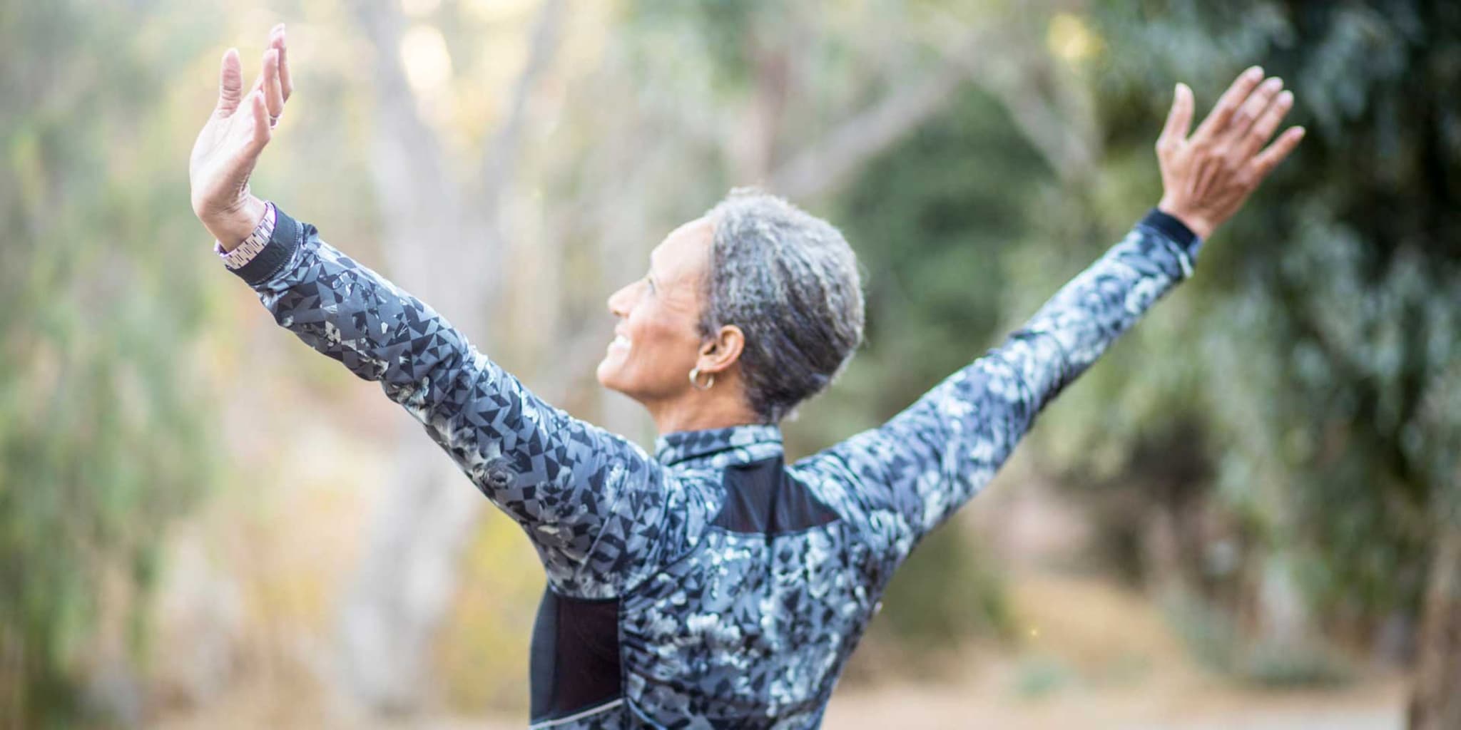 DAK-Rückentraining für Senioren: Ältere Frau in Sportkleidung streckt draußen ihre Arme seitlich aus.
