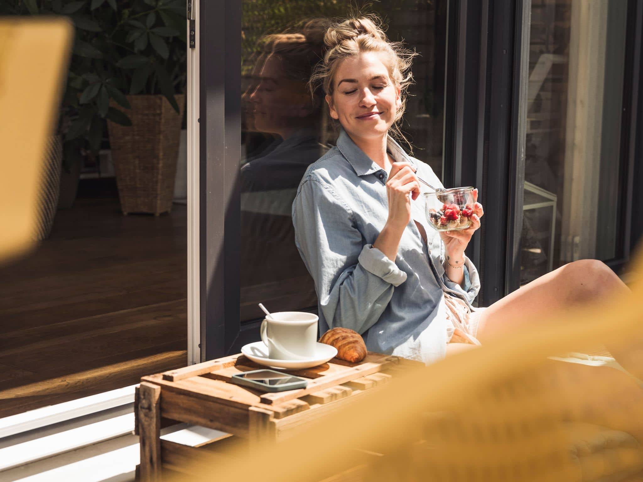 Eine junge, gelassene Frau sitzt auf dem Balkon und isst entspannt Müsli