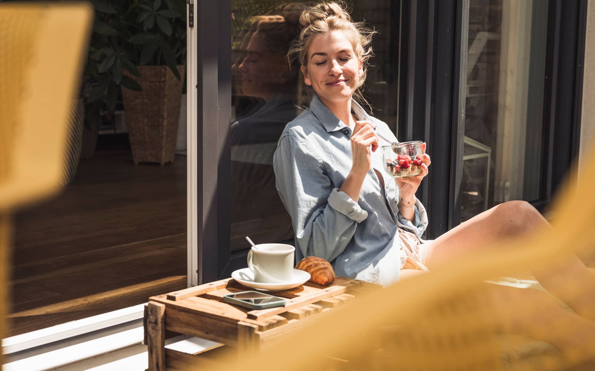 Eine junge, gelassene Frau sitzt auf dem Balkon und isst entspannt Müsli