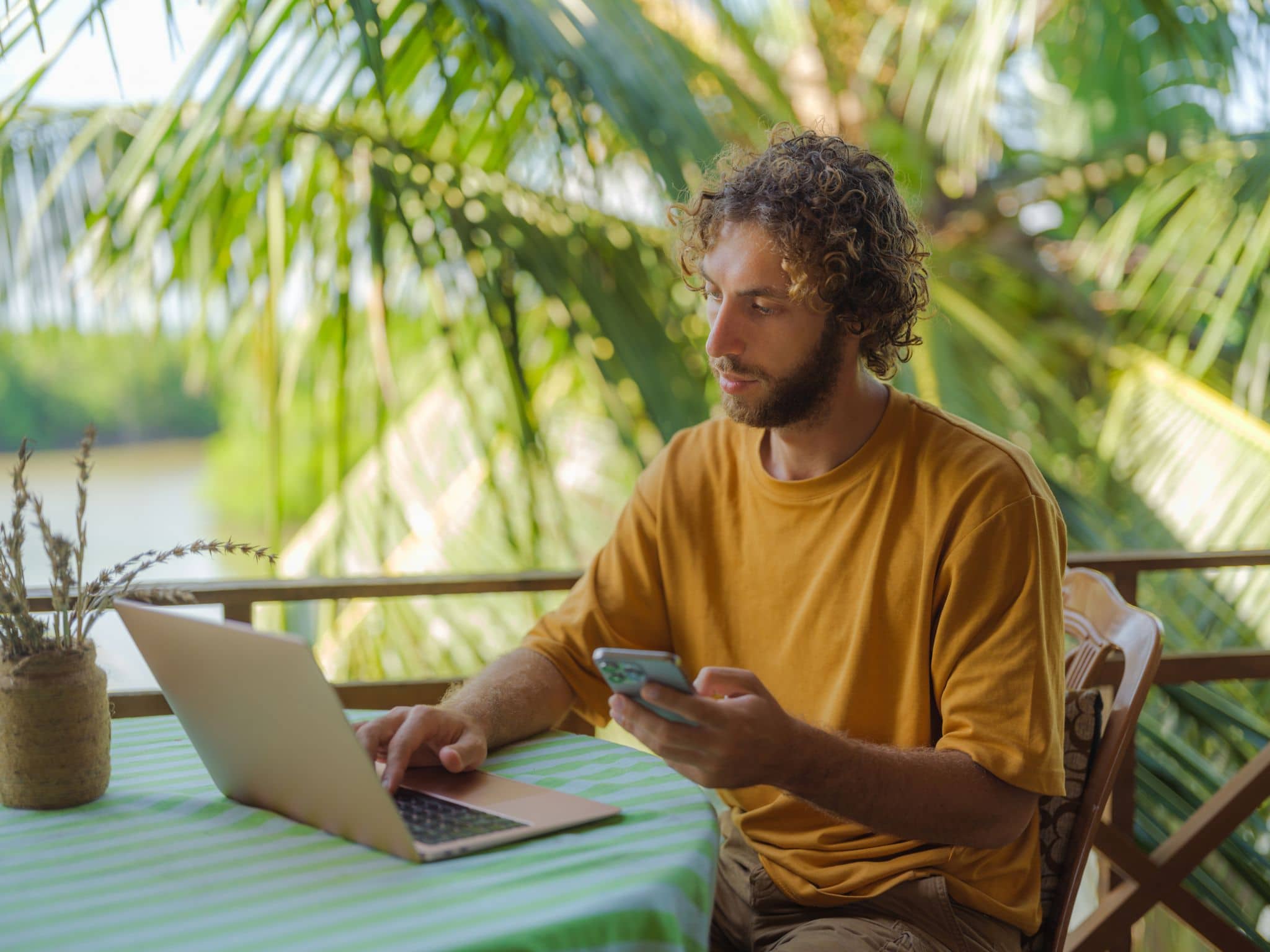 Workation: Mann mit Notebook und Handy am Tisch vor Palmen.