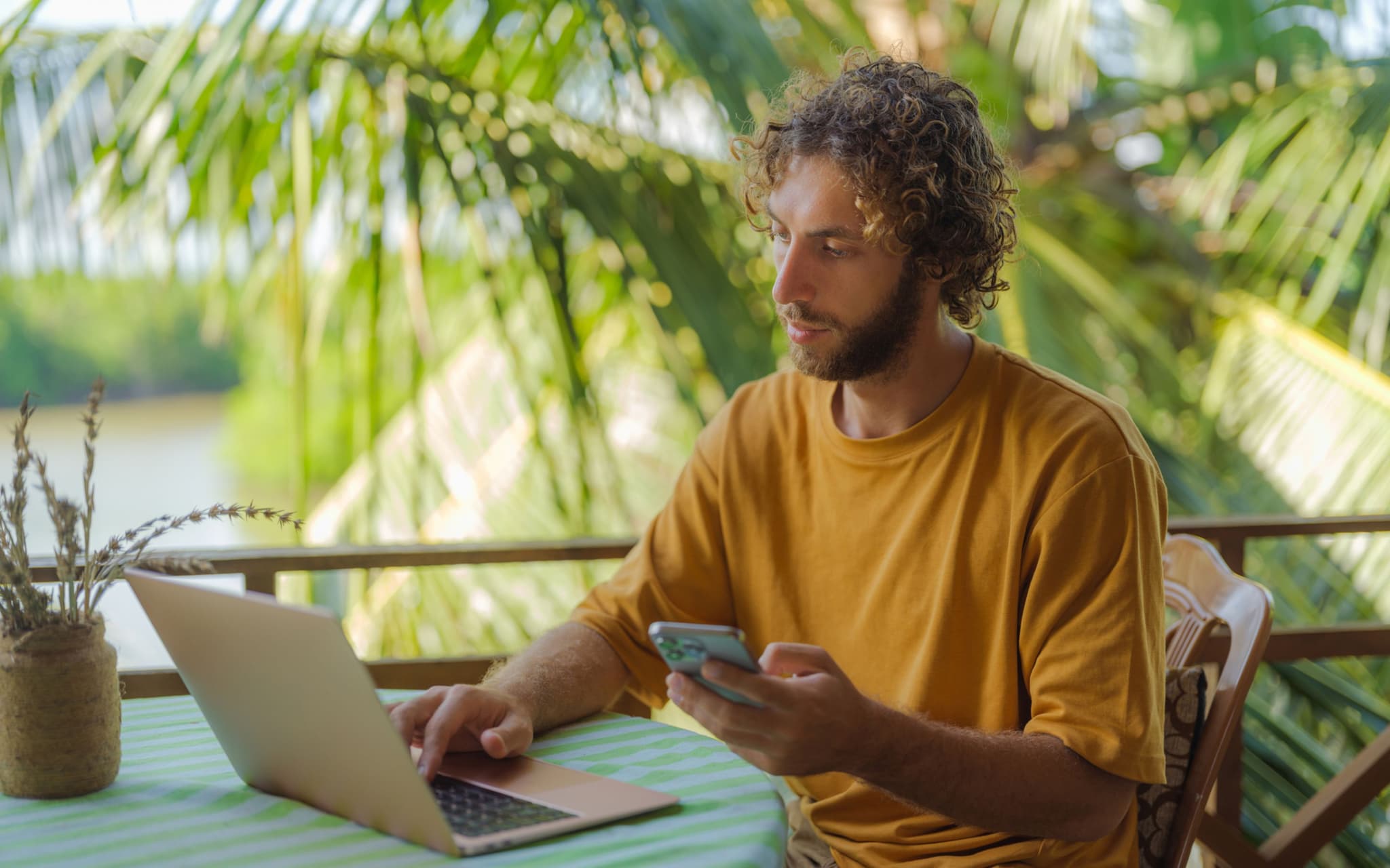 Workation: Mann mit Notebook und Handy am Tisch vor Palmen.