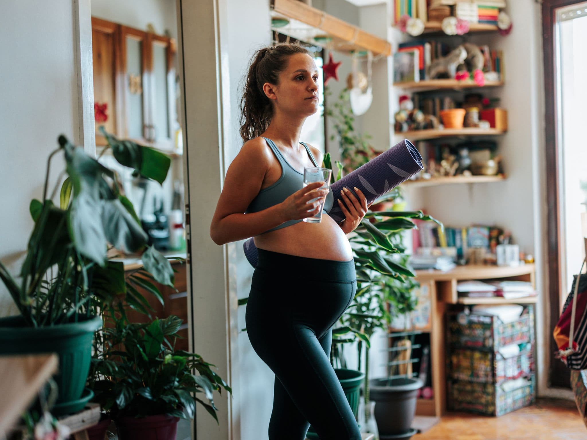 Schwangerschaftsmythen: Eine Schwangere Frau in Sportkleidung trägt eine Gymnastikmatte und trinkt ein Glas Wasser