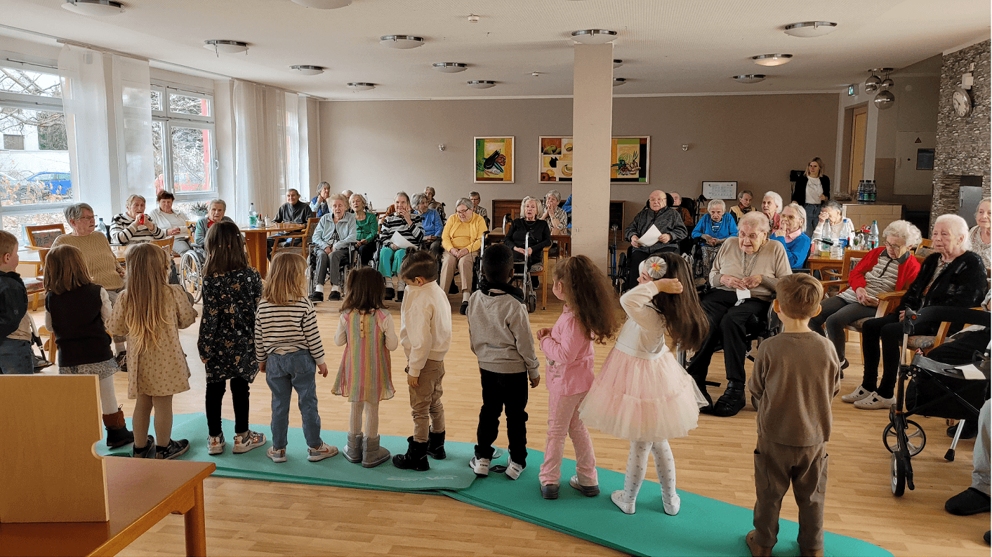 Bild: Gruppenbild ältere Personen Sitzen im Raum und vor ihnen stehen mehrere Kinder