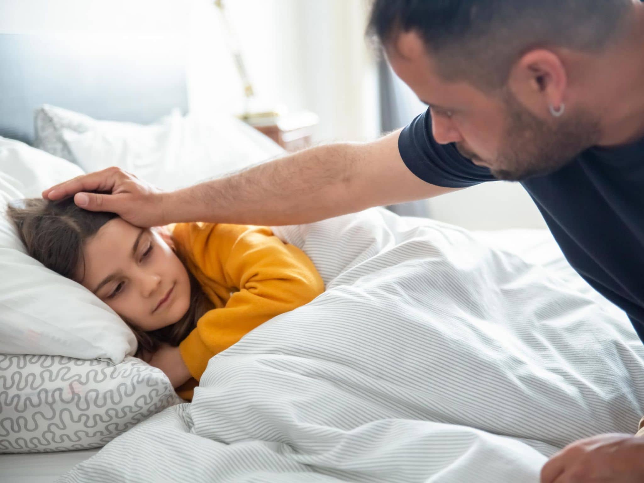 Scharlach: Ein Vater fasst seiner kranken, im Bett liegenden Tochter an die Stirn.