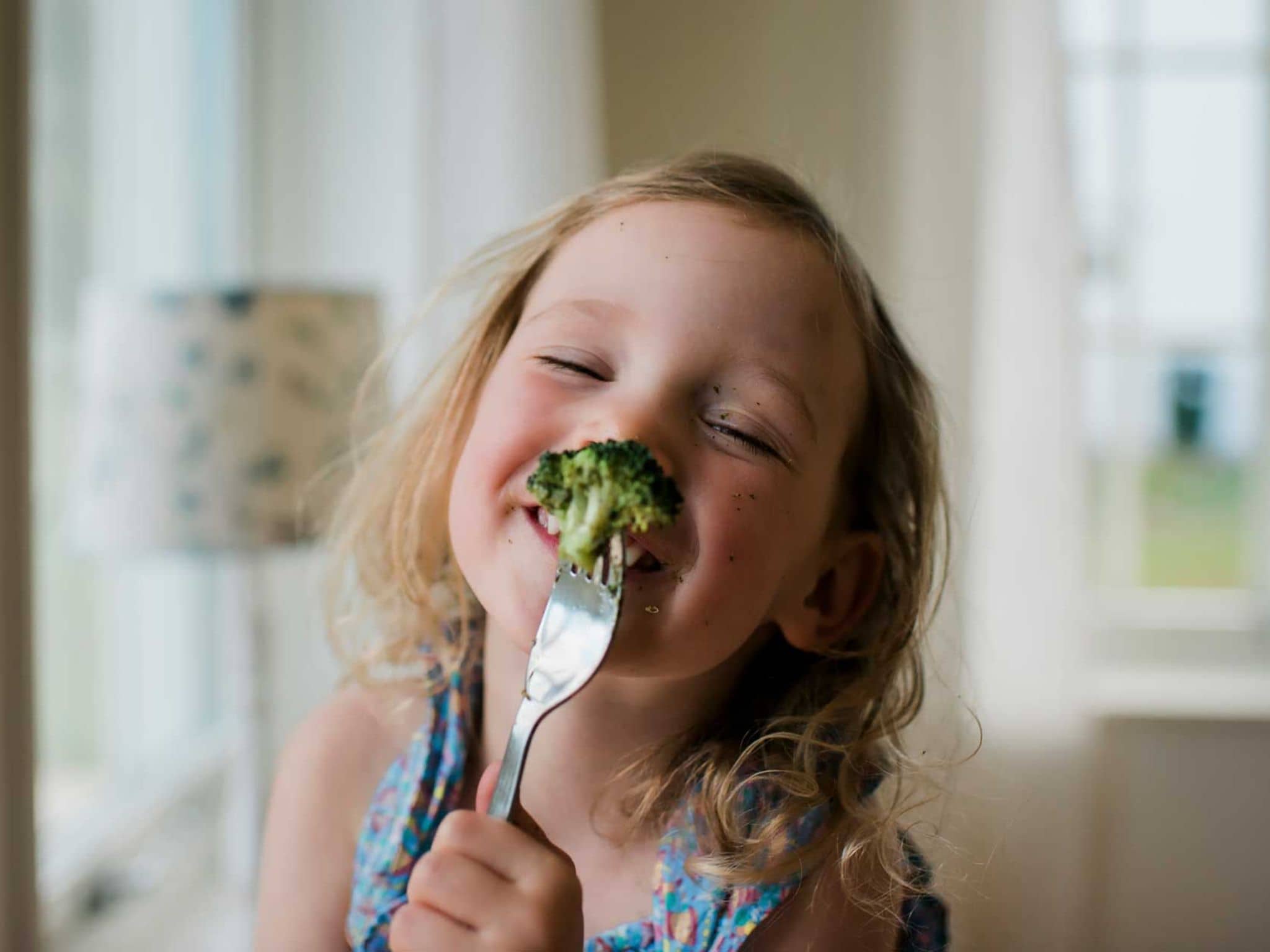 Kohl ist gesund: Lachendes kleines Mädchen hält Gabel mit aufgespießten Brokkoli vors Gesicht.