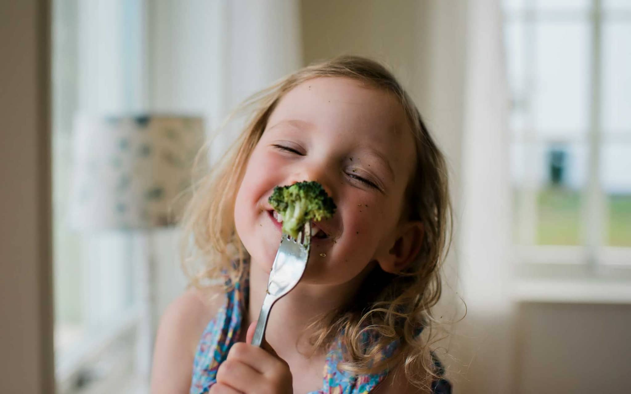 Kohl ist gesund: Lachendes kleines Mädchen hält Gabel mit aufgespießten Brokkoli vors Gesicht.