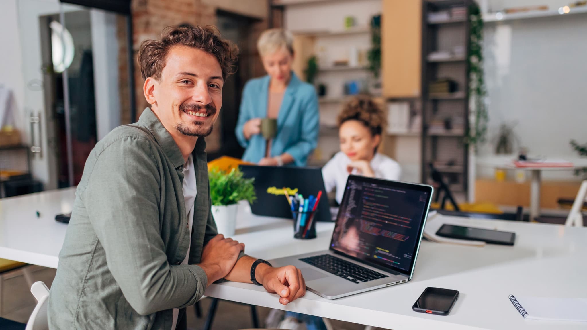 Nasmejani muškarac sedi sa kolegama za konferencijskim stolom ispred laptopa i gleda u kameru.
