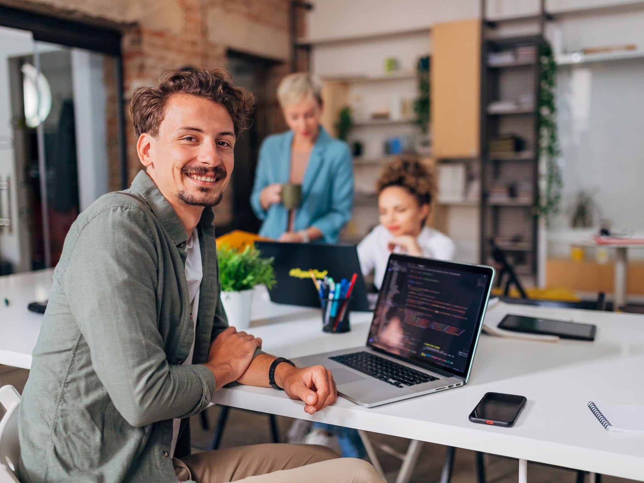 Nasmejani muškarac sedi sa kolegama za konferencijskim stolom ispred laptopa i gleda u kameru.