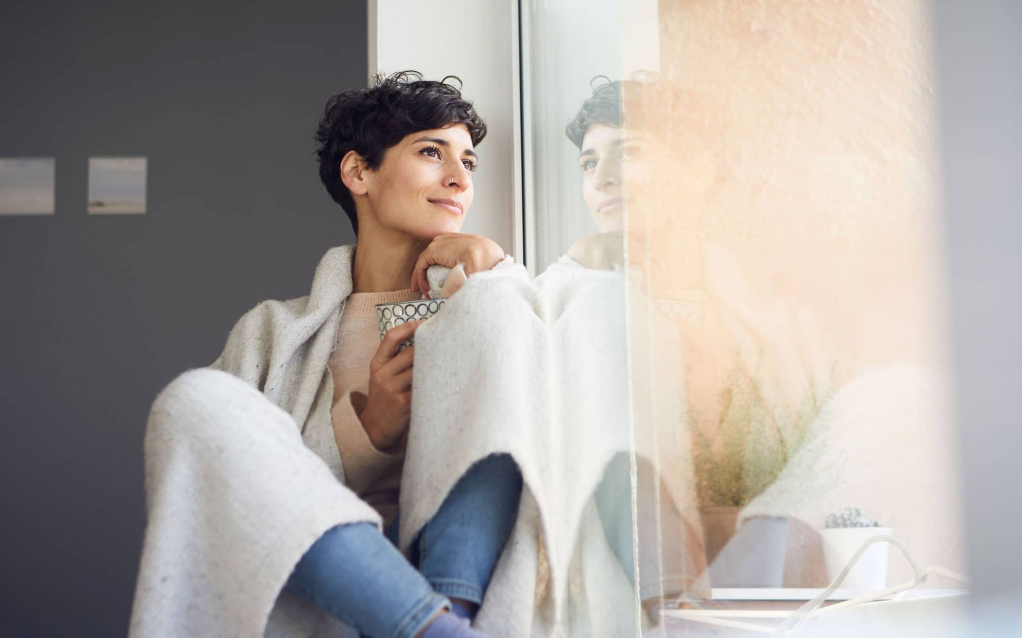 Junge Frau sitzt in eine Wolldecke gehüllt am Fenster und schaut hinaus.