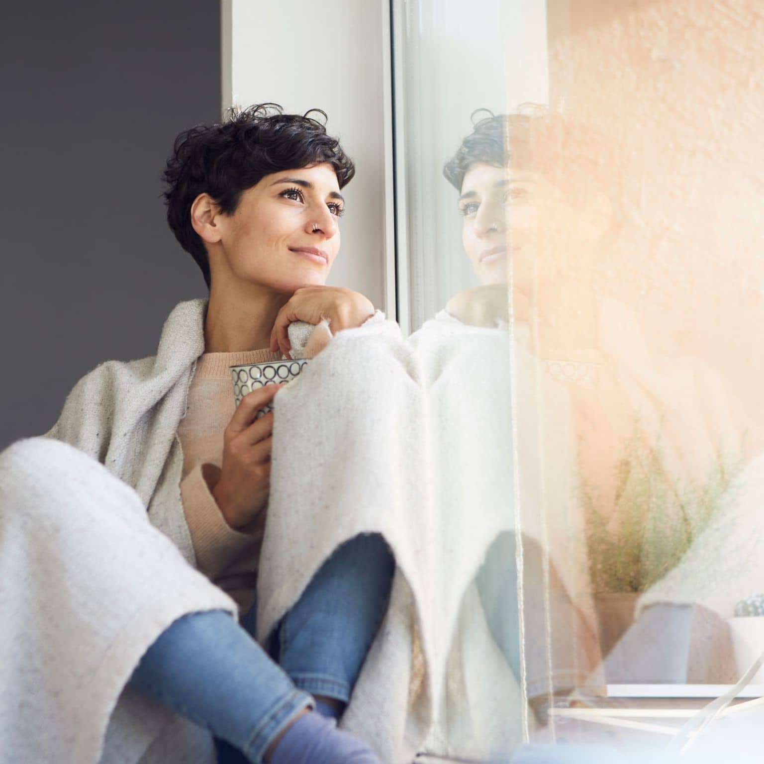 Junge Frau sitzt in eine Wolldecke gehüllt am Fenster und schaut hinaus.