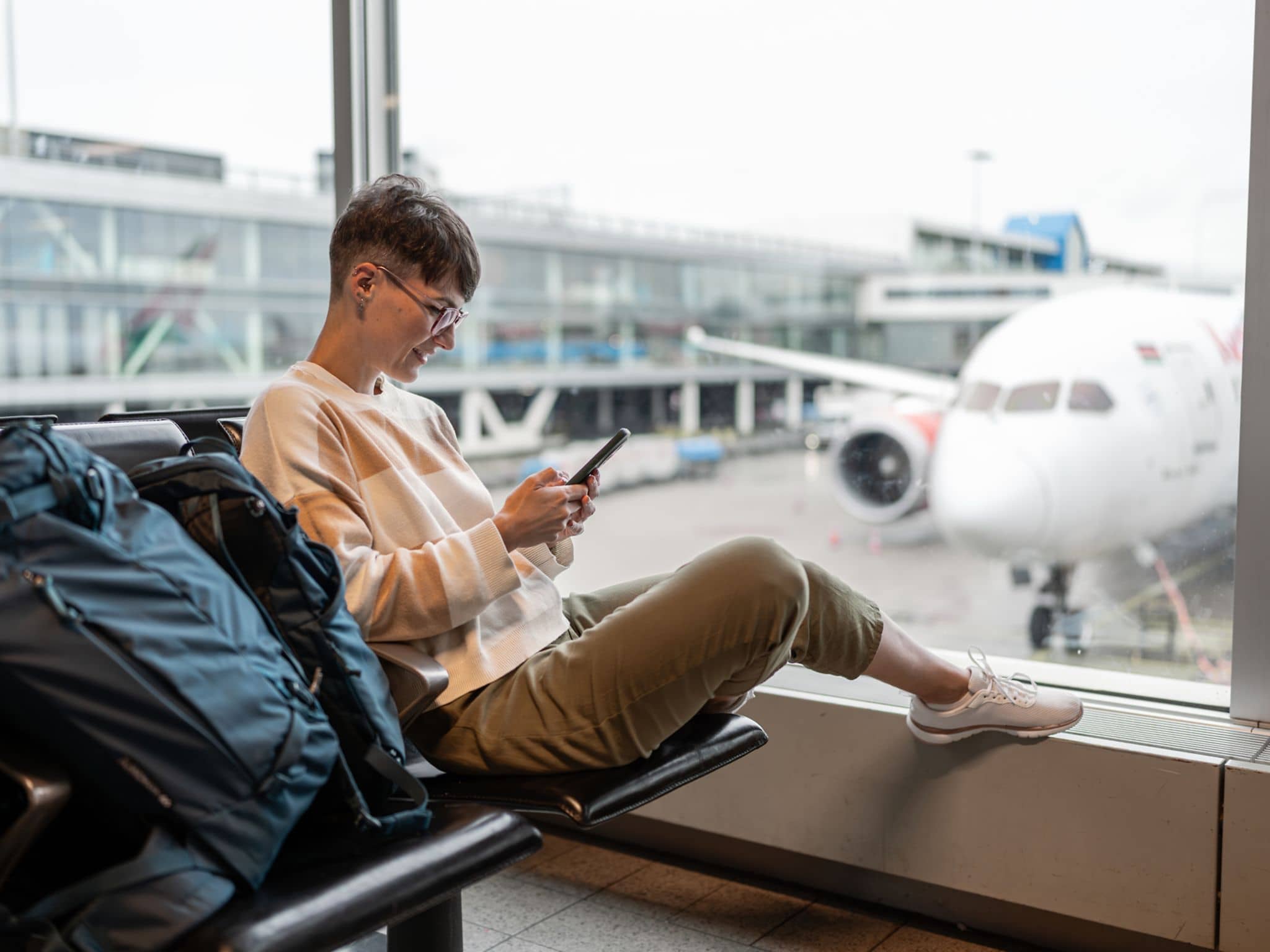 Frau sitzt im Abflug-Bereich eines Flughafens und schaut auf ein Handy.