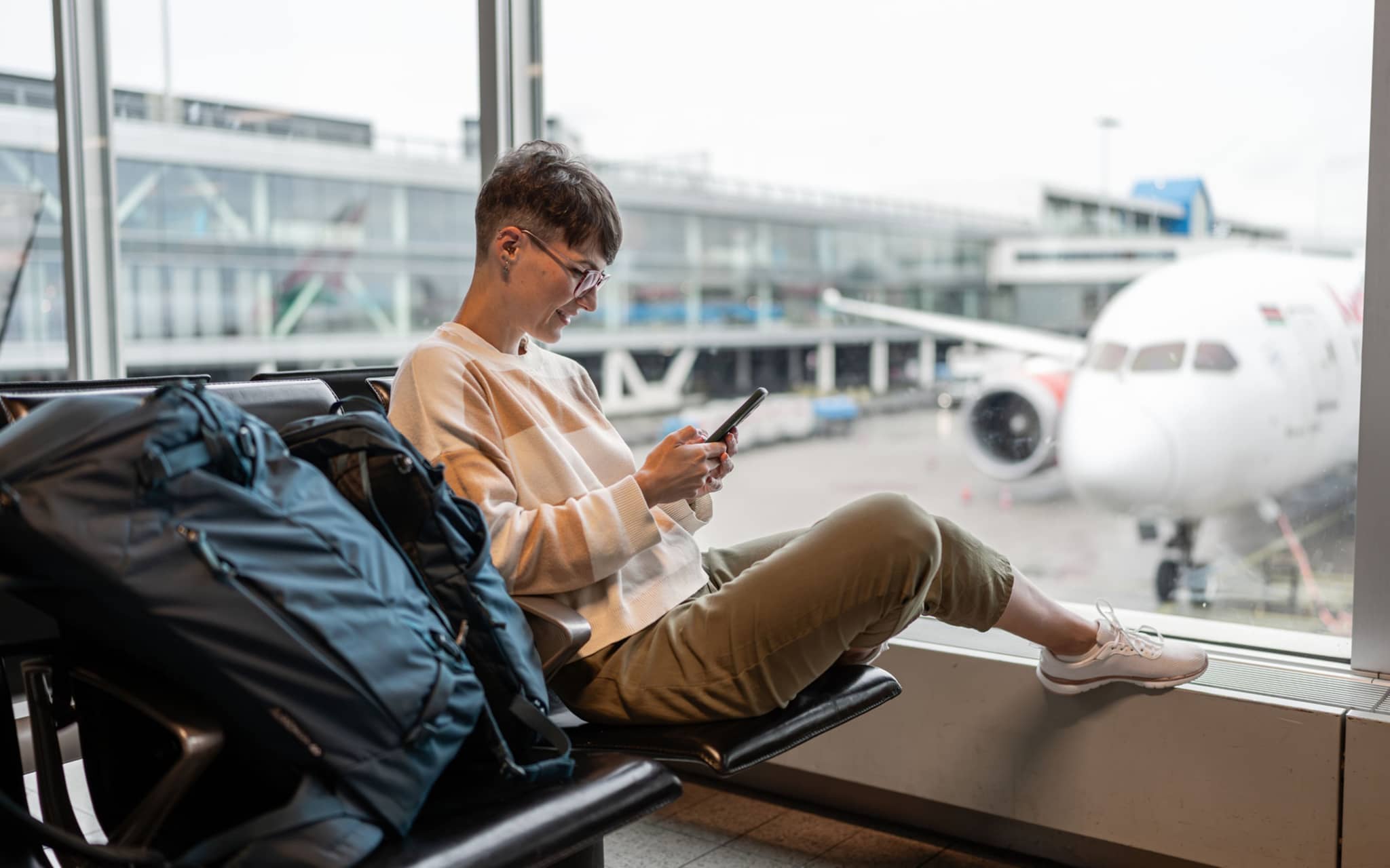 Frau sitzt im Abflug-Bereich eines Flughafens und schaut auf ein Handy.