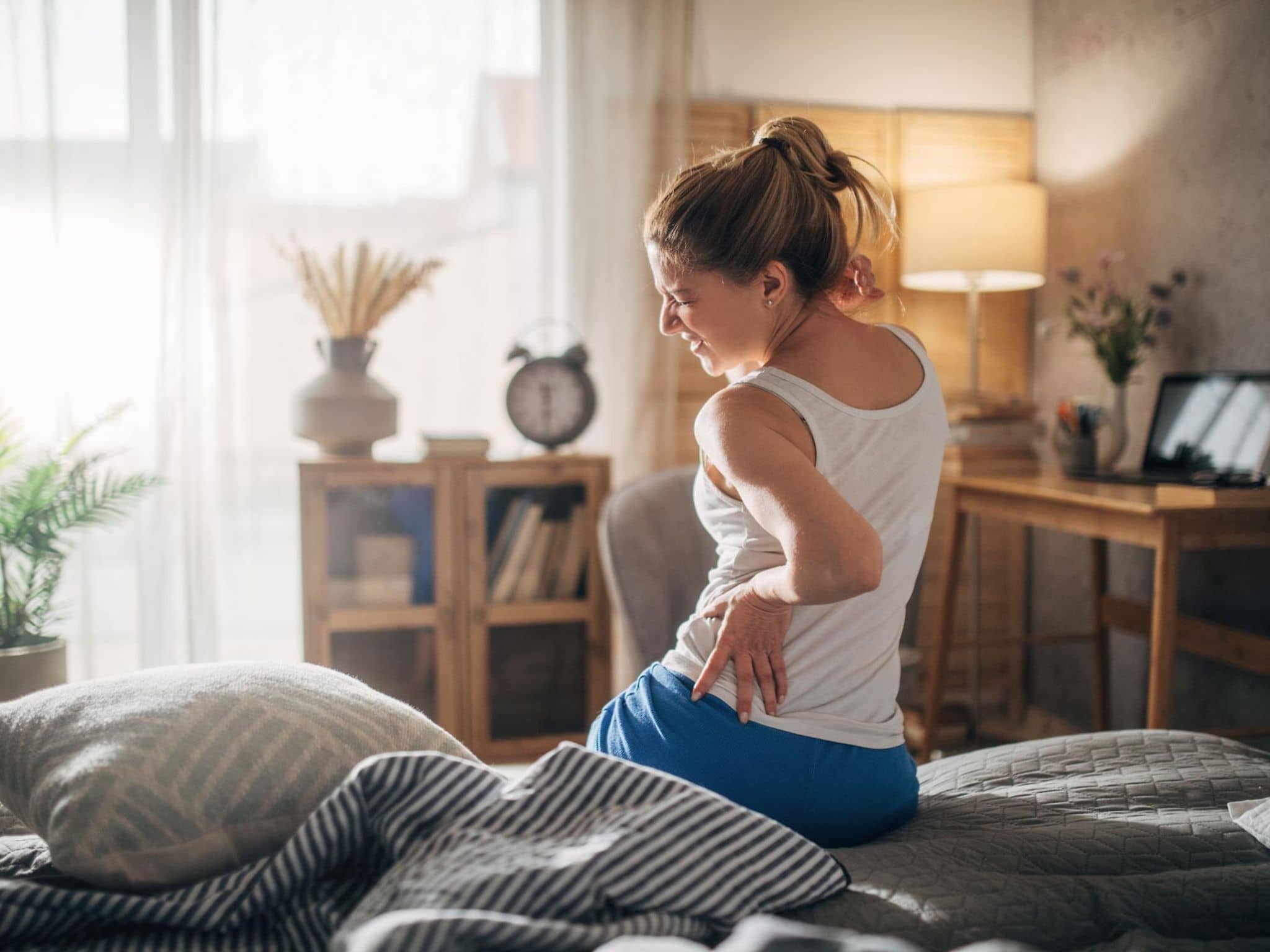 Junge Frau sitzt auf der Bettkante und fasst sich mit schmerzverzerrtem Gesicht an den Rücken.