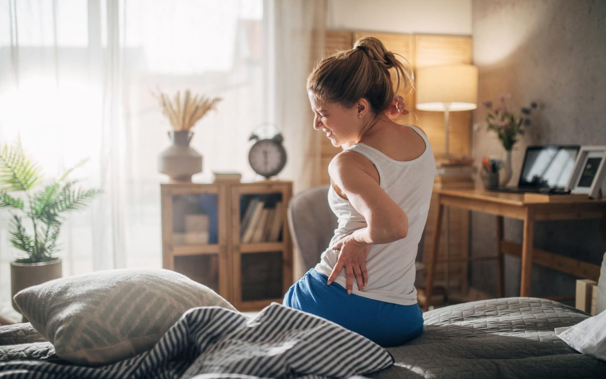 Junge Frau sitzt auf der Bettkante und fasst sich mit schmerzverzerrtem Gesicht an den Rücken.