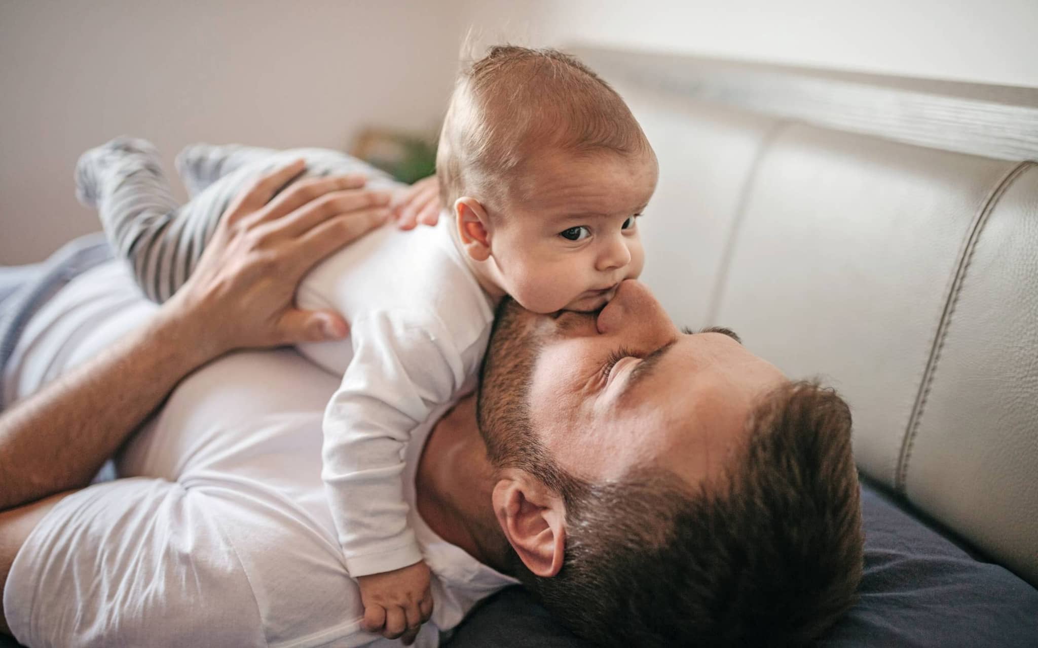Bild: Junger Vater liegt auf dem Sofa und gibt seinem Baby einen Kuss.