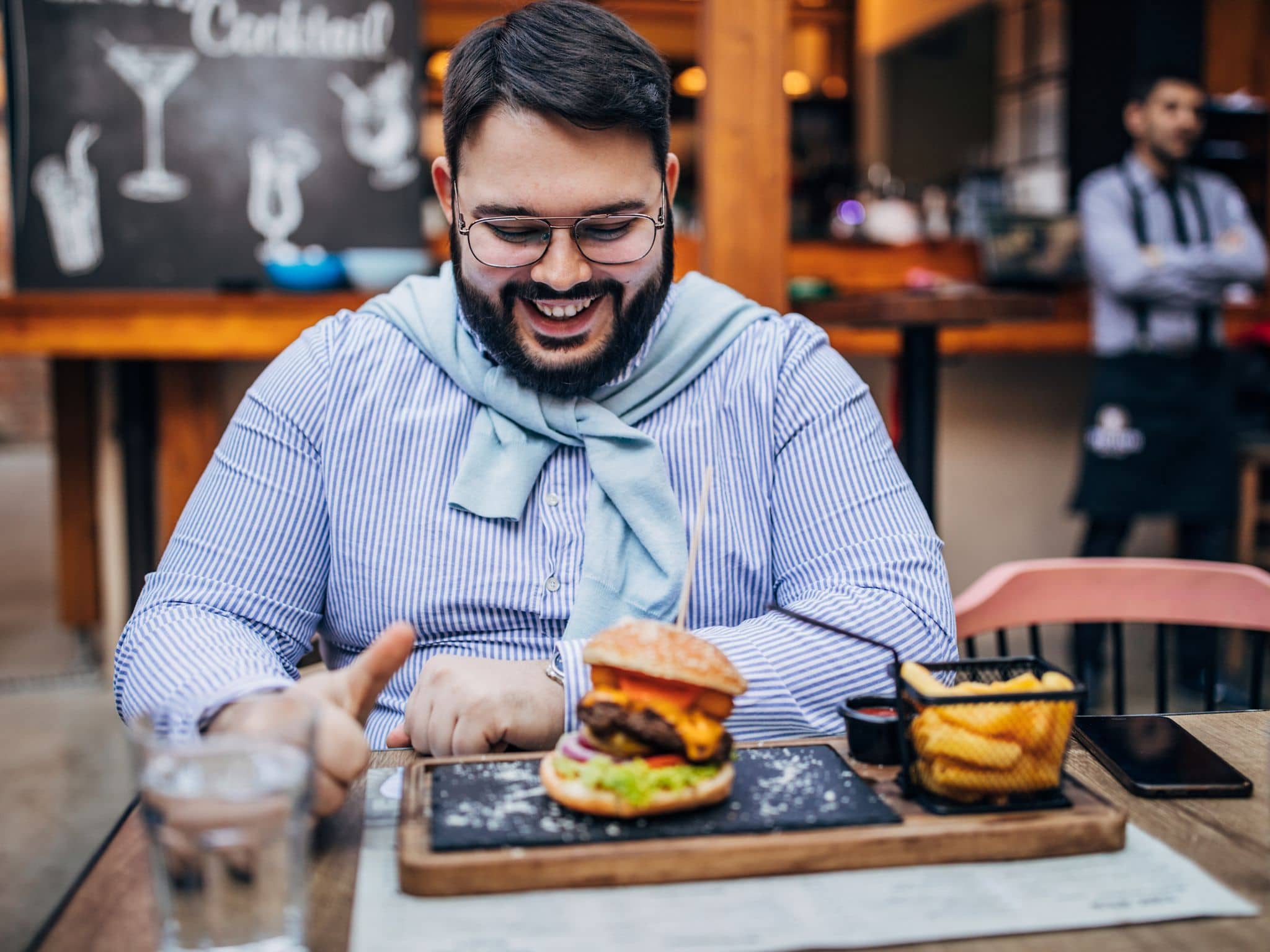 Bild: Korpulenter Mann sitzt in einem Restaurant und freut sich auf seinen Hamburger.