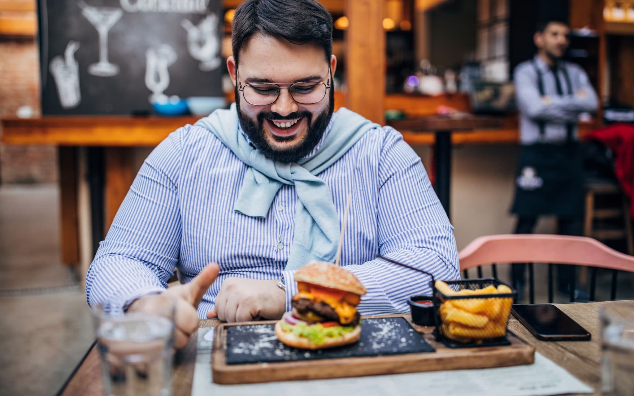 Bild: Korpulenter Mann sitzt in einem Restaurant und freut sich auf seinen Hamburger.