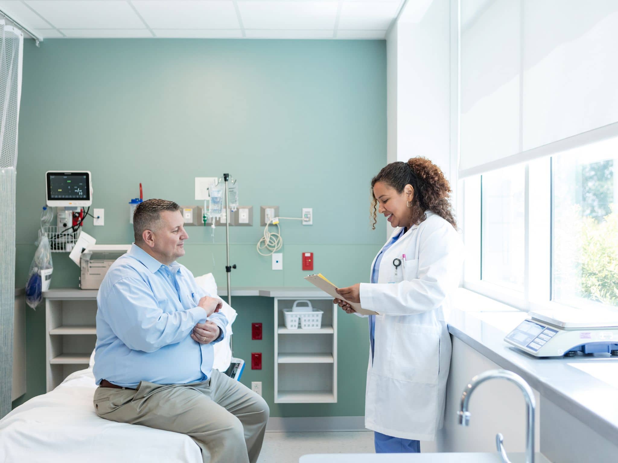 Bild: Mann mit Adipositas sitzt auf einem Krankenbett während die Ärztin einen Bericht liest.