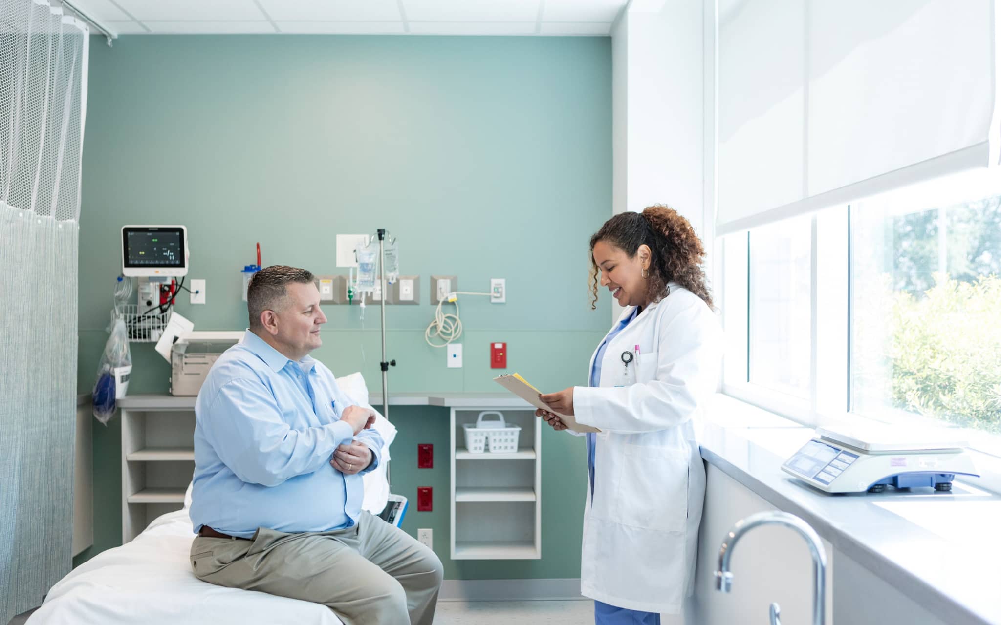 Bild: Mann mit Adipositas sitzt auf einem Krankenbett während die Ärztin einen Bericht liest.