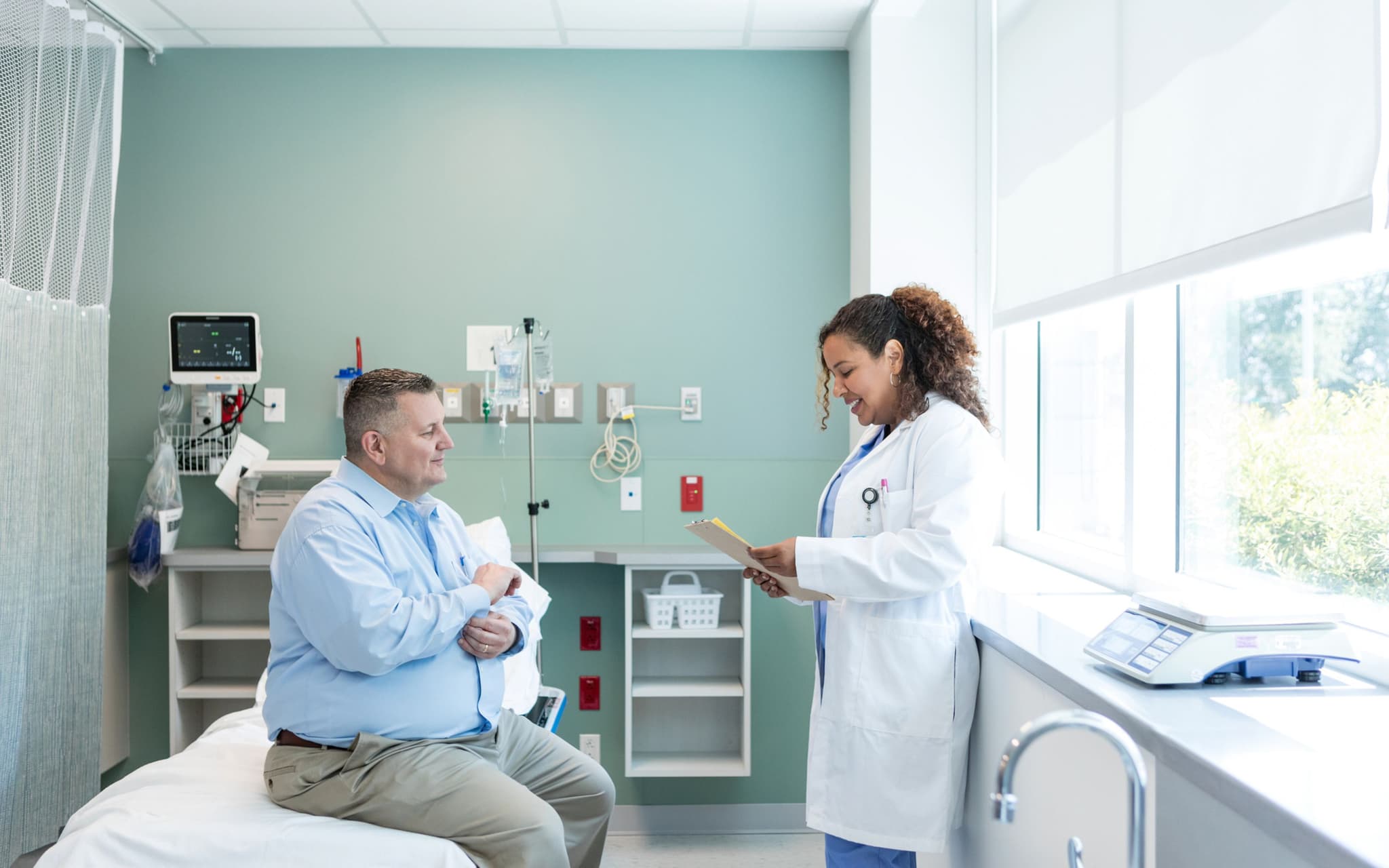 Bild: Mann mit Adipositas sitzt auf einem Krankenbett während die Ärztin einen Bericht liest.