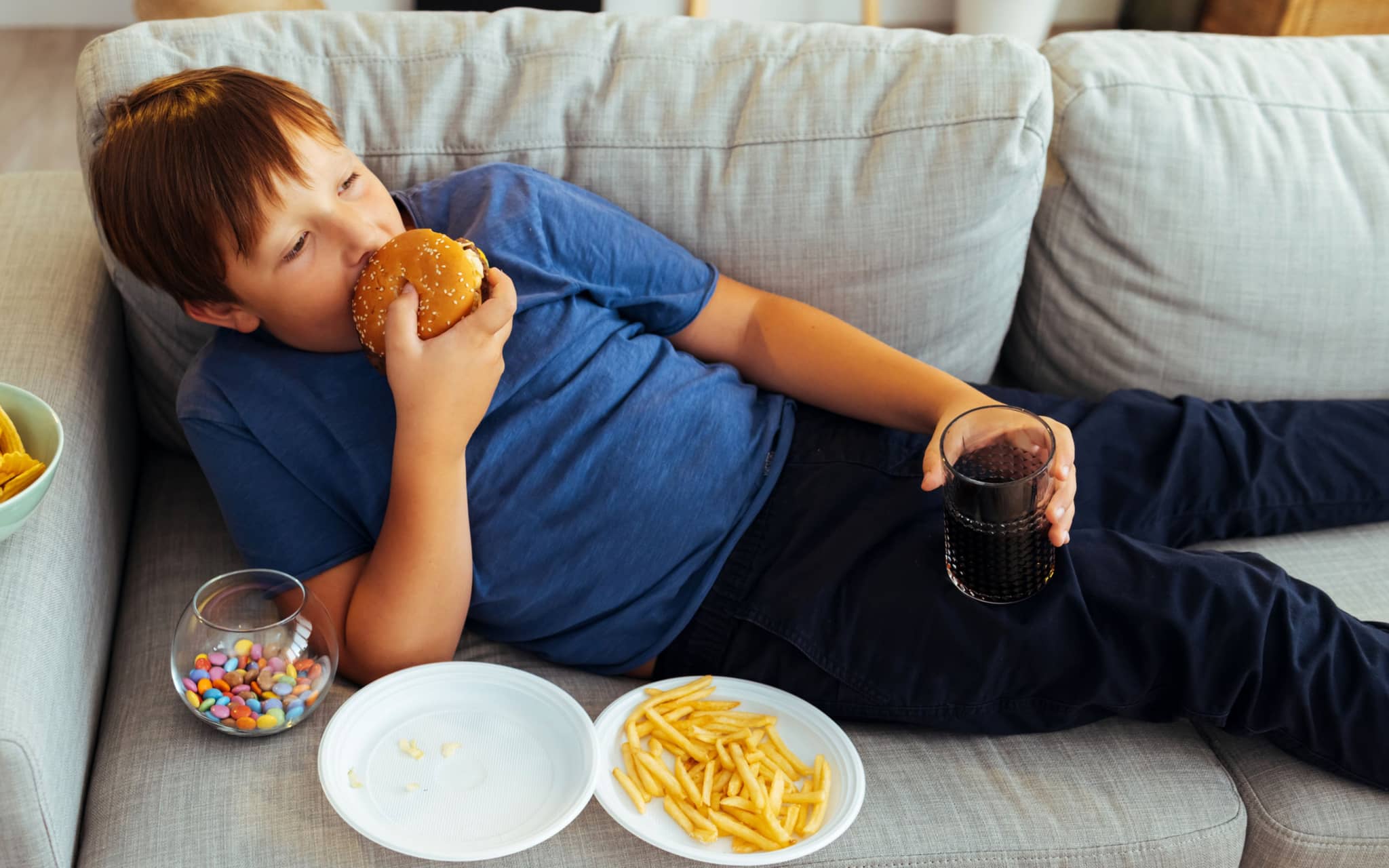 Adipositas: Ein kleiner Junge liegt auf dem Sofa und isst einen Hamburger und Pommes Frites.