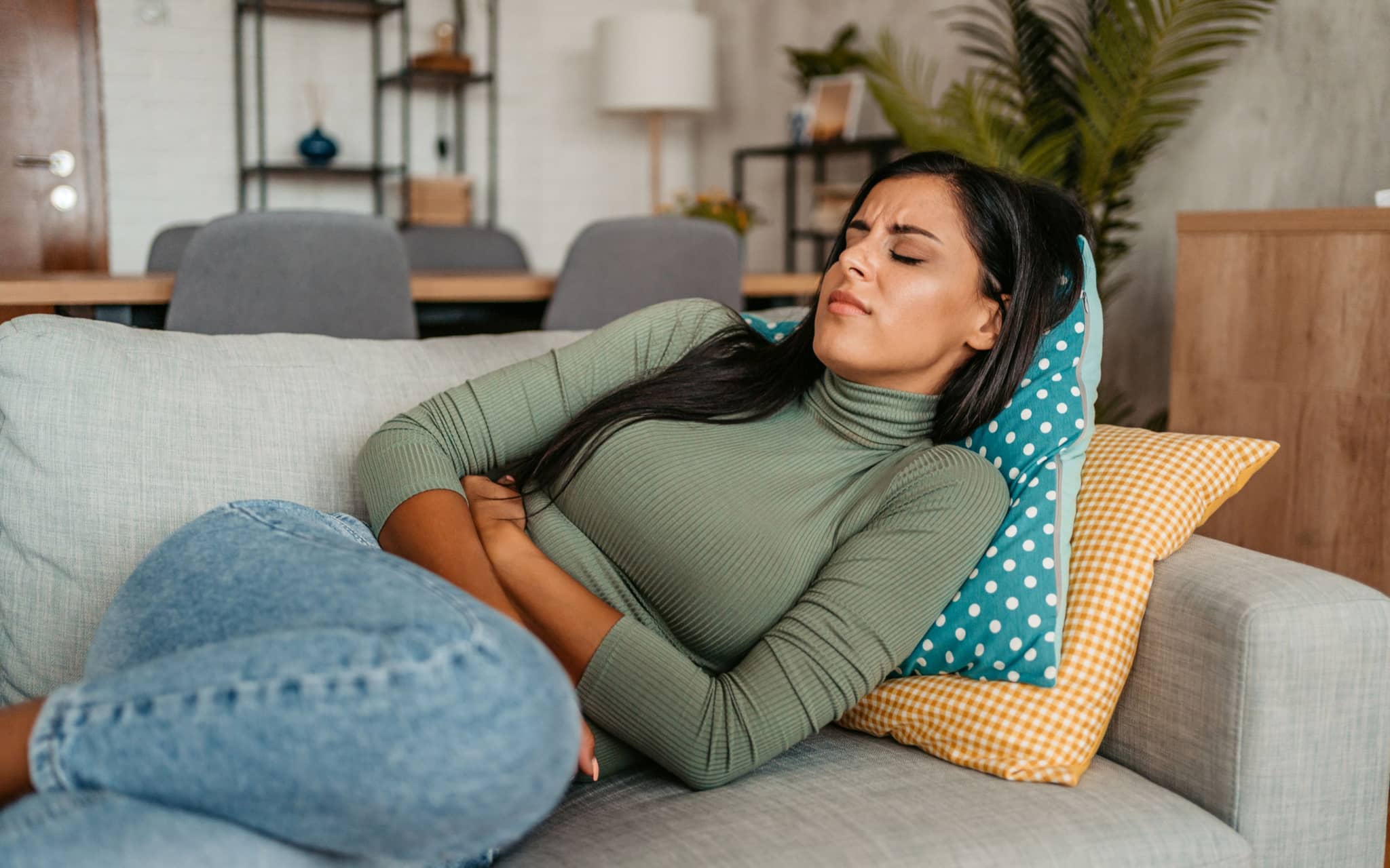 Zöliakie: Junge Frau liegt auf ihrem Sofa und verspürt Schmerzen im Bauch