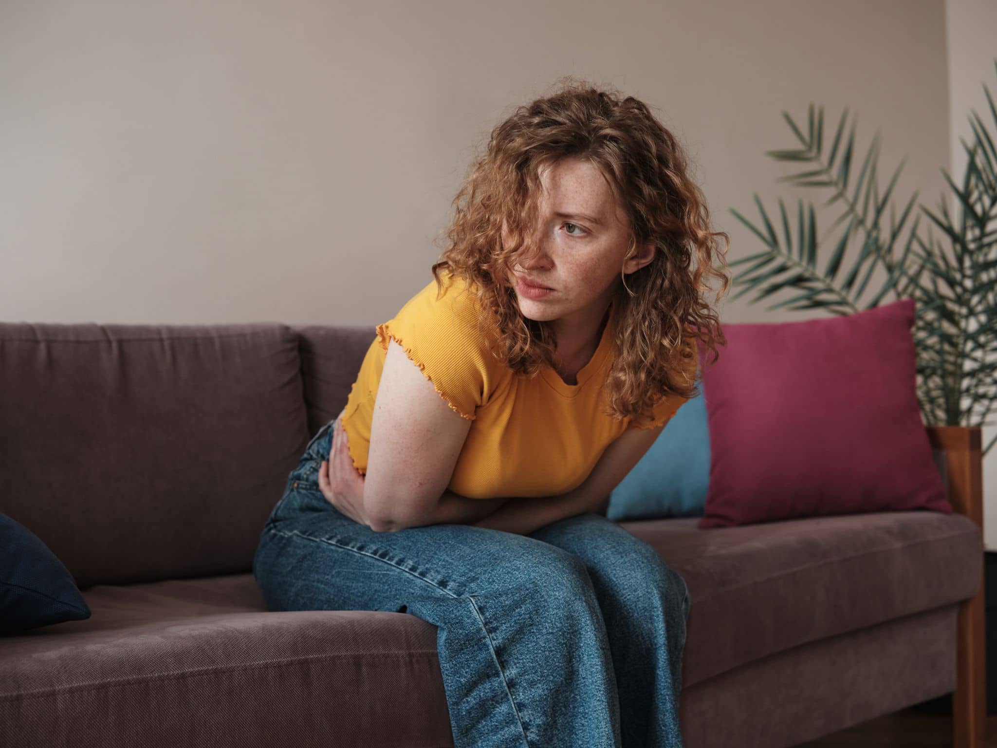 Histaminintoleranz: Junge Frau sitzt nach vorn gebeugt mit Bauchschmerzen auf ihrem Sofa