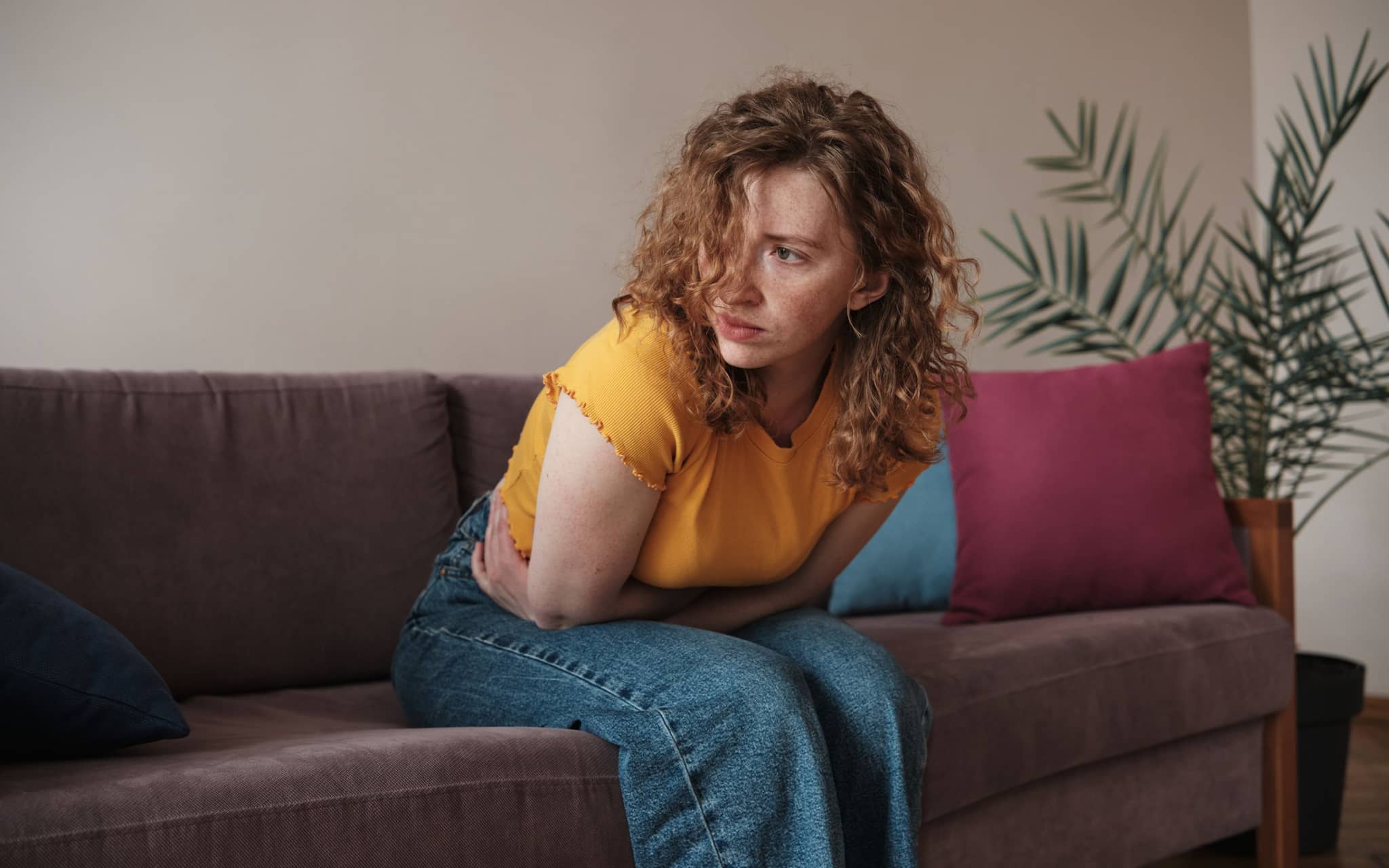 Histaminintoleranz: Junge Frau sitzt nach vorn gebeugt mit Bauchschmerzen auf ihrem Sofa