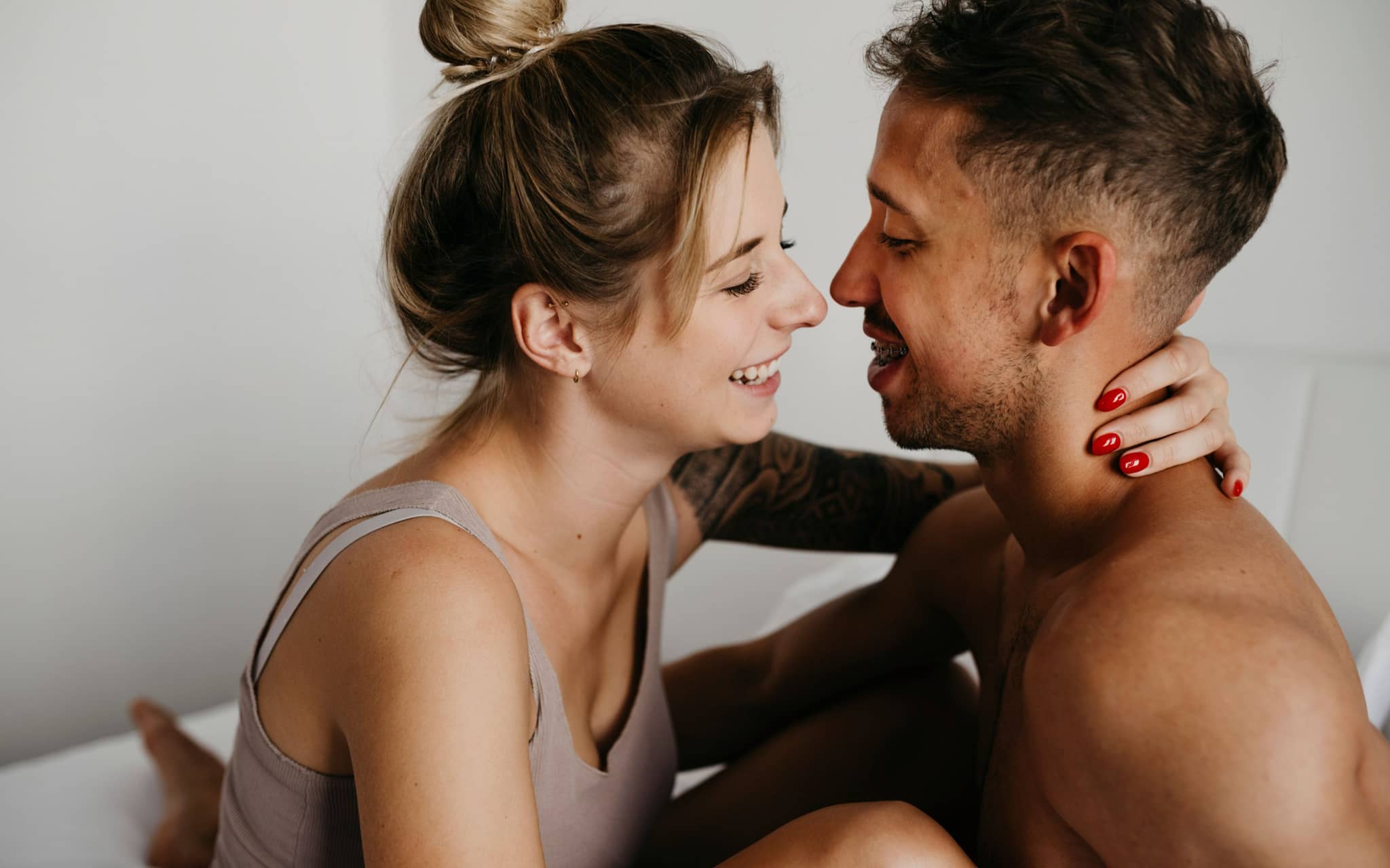 Blowjob: Eine junge Frau und ein junger Mann sitzen in Unterwäsche im Bett, lächeln sich an und umarmen sich.