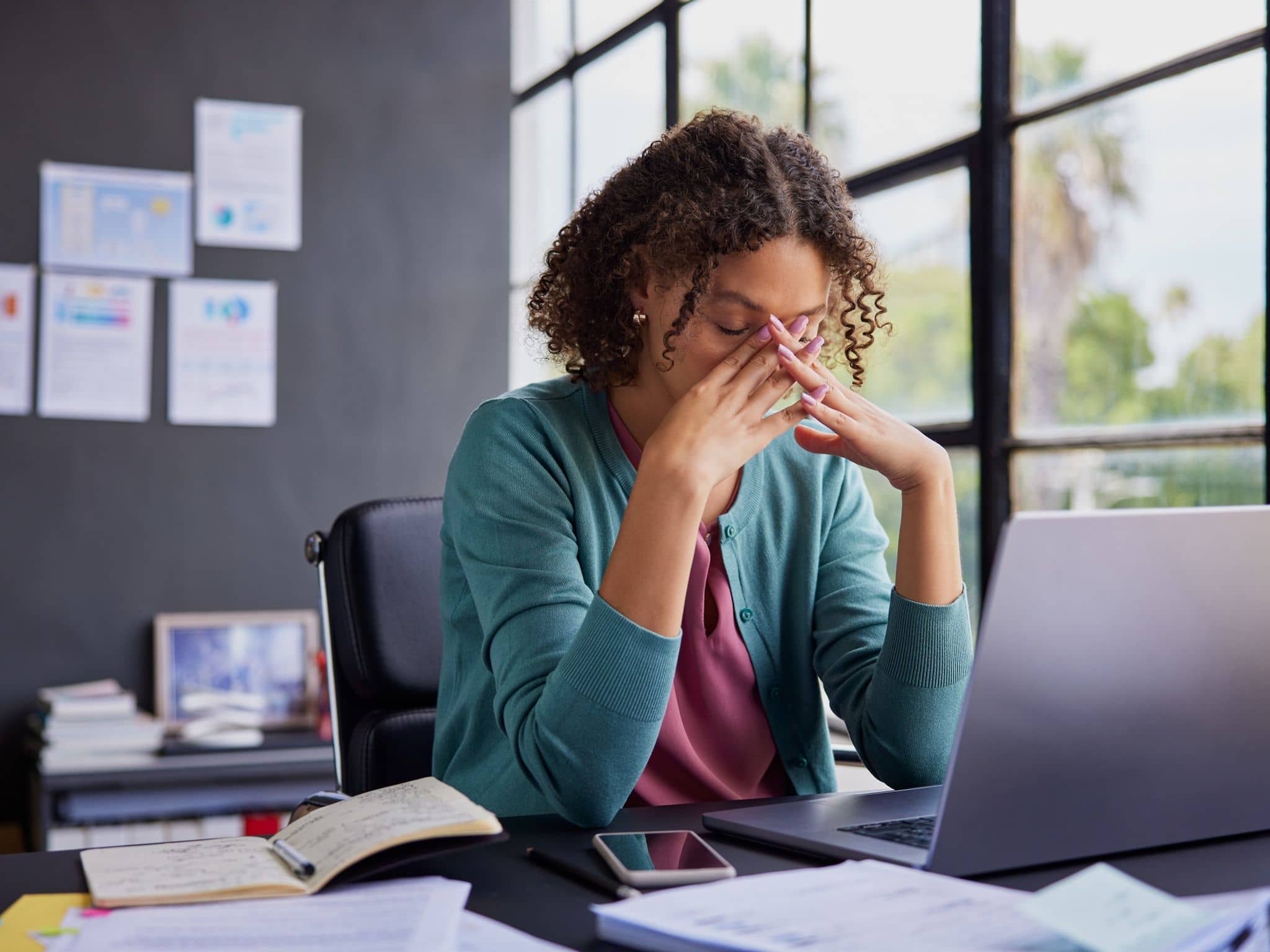 Psychreport: Erschöpft wirkende Frau sitzt vor einem Notebook und drückt sich die Finger vor die verschlossenen Augen..