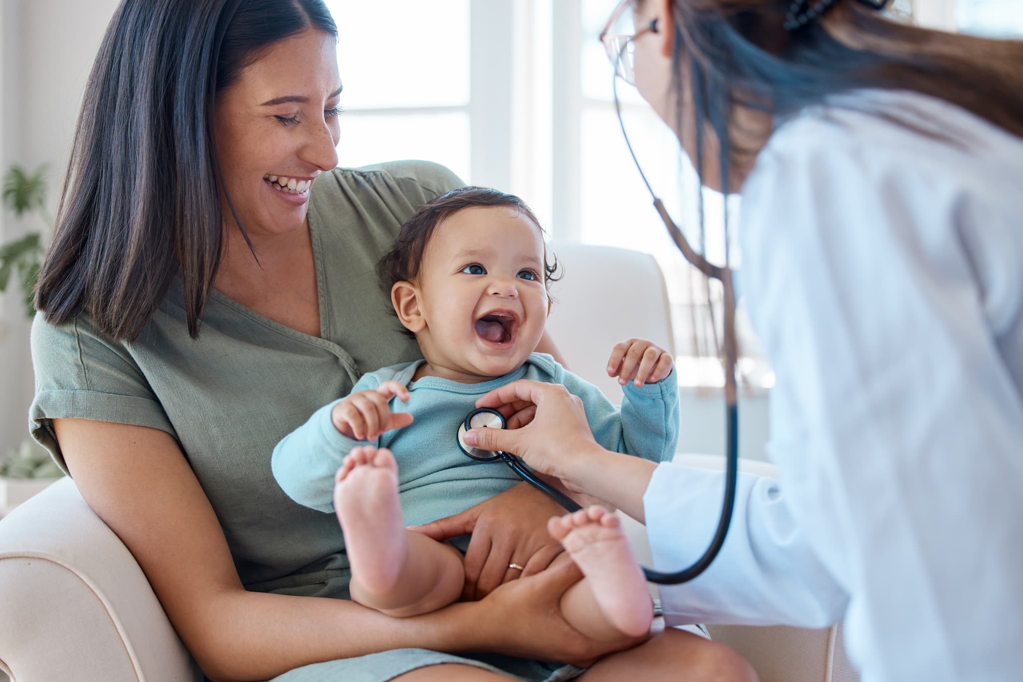 Kindervorsorge: Junge Mutter hält ihr Baby freudig auf dem Arm, während eine Ärztin es kardiologisch untersucht