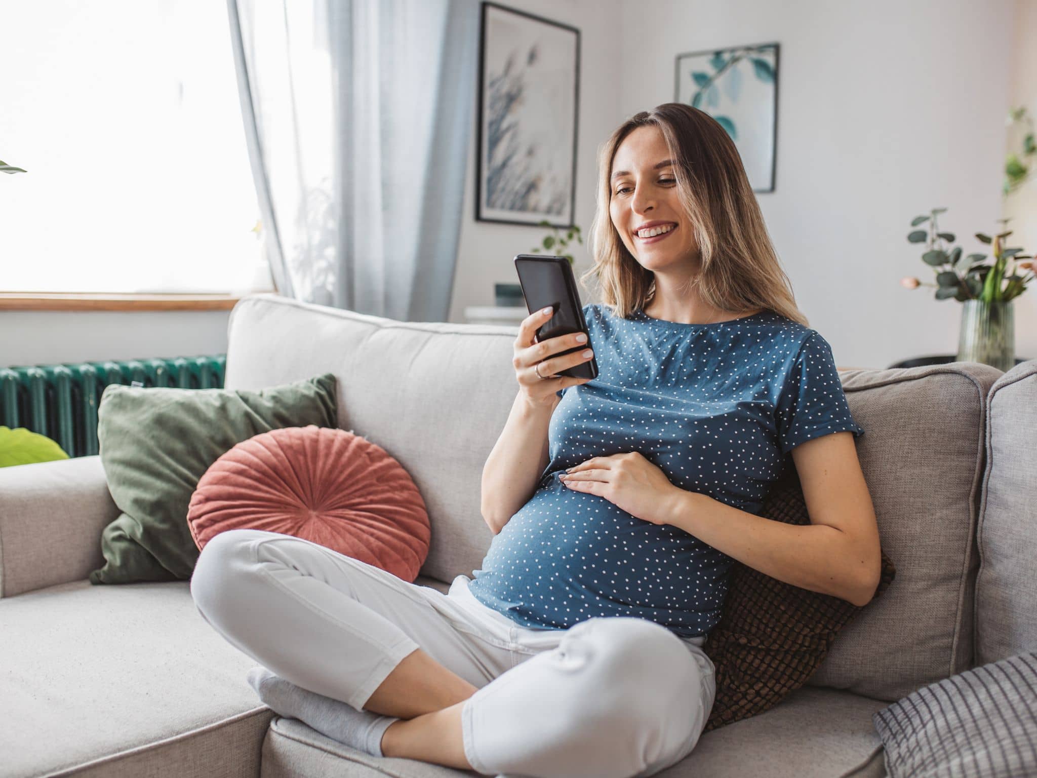 Symbolbild BabyCare App: Eine Schwangere sitzt auf dem Sofa und liest auf ihrem Smartphone