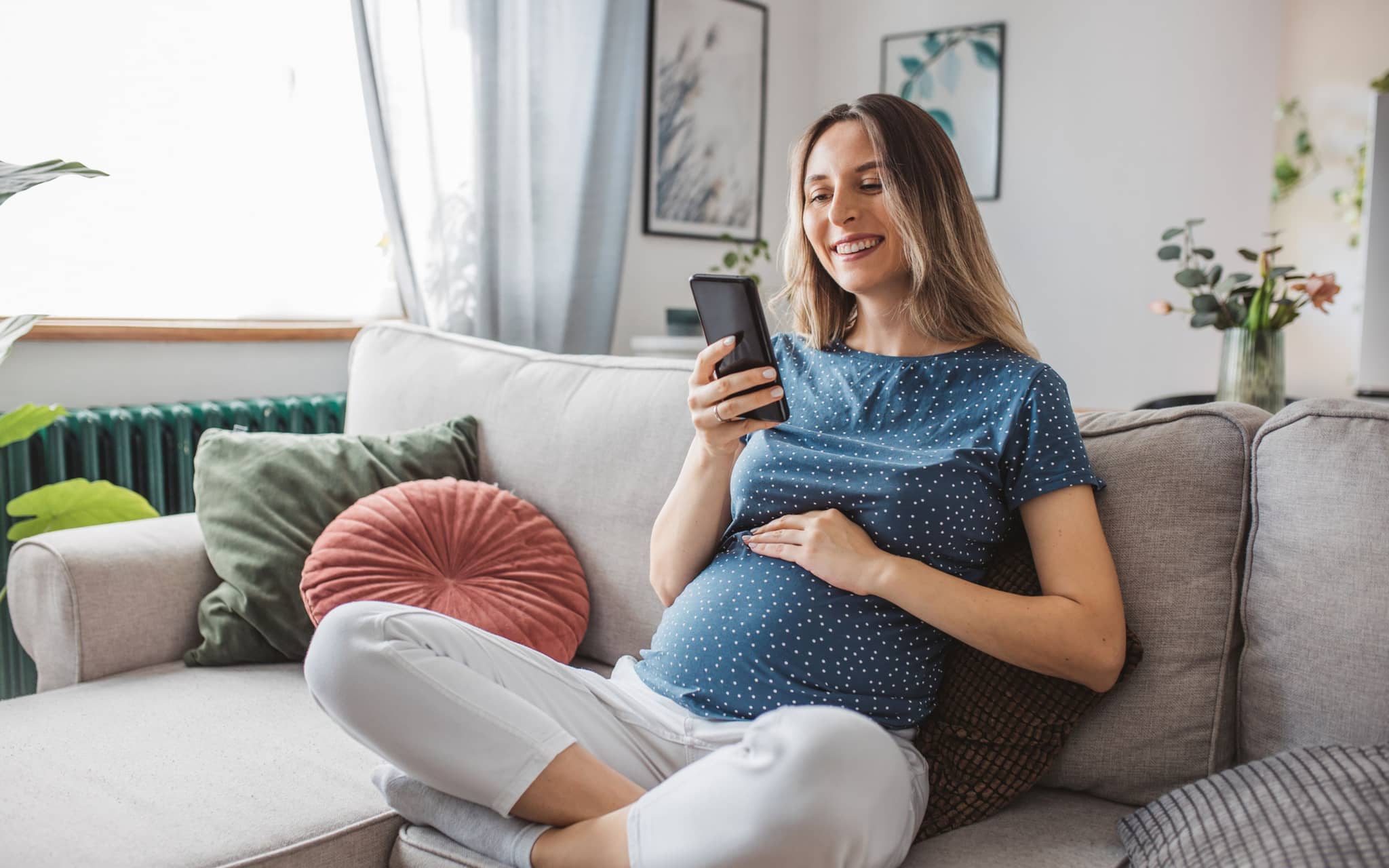 Symbolbild BabyCare App: Eine Schwangere sitzt auf dem Sofa und liest auf ihrem Smartphone