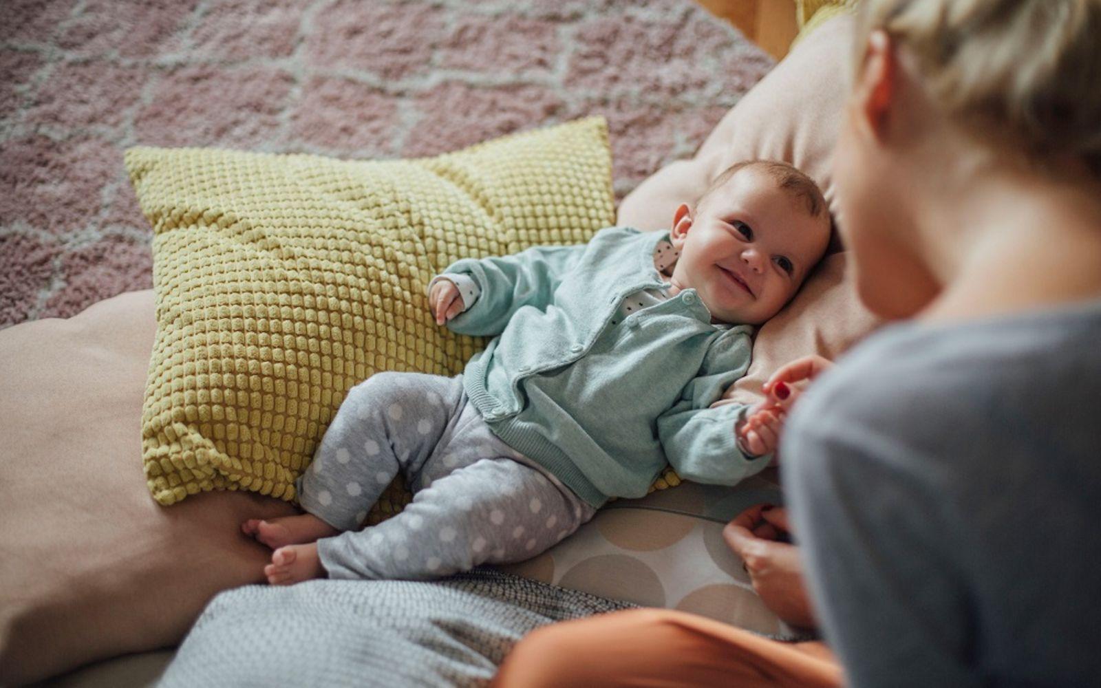 DAK Willkommen Baby: Lächelndes Baby liegt auf Kissen und schaut zu einer Frau hoch.